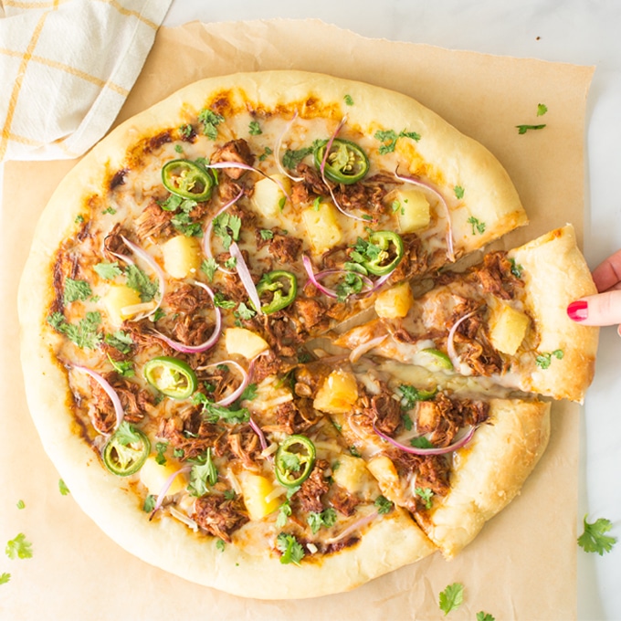 hand grabbing slice pf jackfruit pizza with bbq sauce, pineapple, red onions, jalapenos, and cilantro