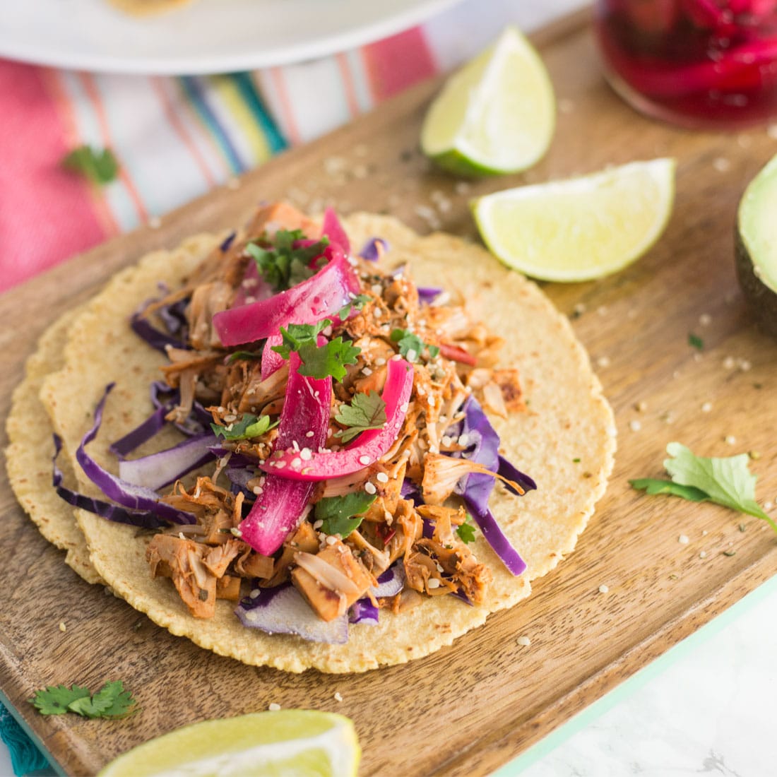 vegan jackfruit pulled pork tacos on wooden board with red onions, lime wedges, cilantro, and purple cabbage