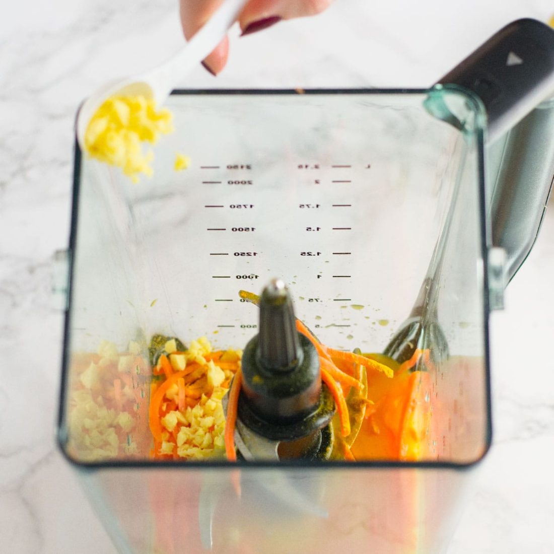 pouring ginger into blender of orange juice, carrots, and turmeric