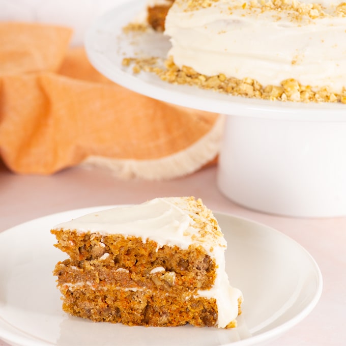 slice of vegan carrot cake with whole cake on a cake stand in the background