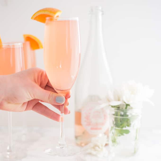 hand holding champagne flute of mimosa with champagne and flowers in background