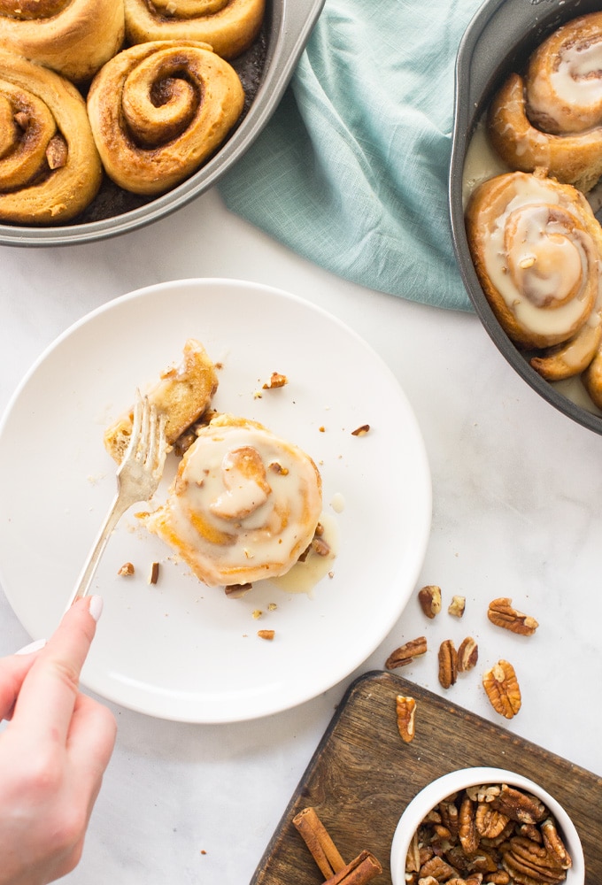 fork eating cinnamon roll with pans of cinnamon rolls, pecans, and cinnamon