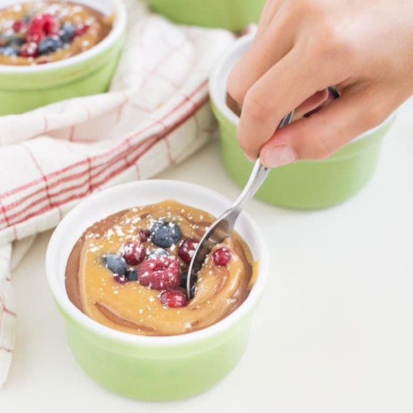 hand dipping spoon into chocolate mousse in green ramekin with caramel, fresh berries, and powdered sugar