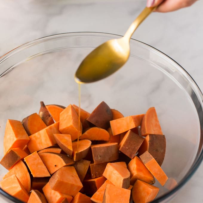 gold spoon pouring olive oil into mixing bowl with diced sweet potatoes
