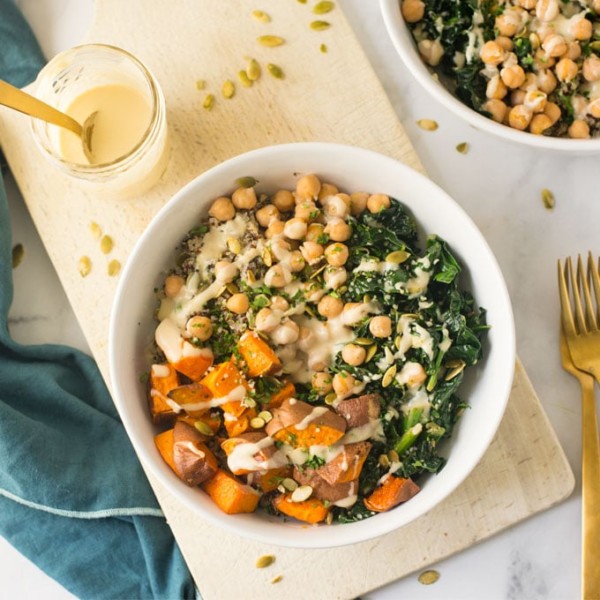 vegan quinoa bowl with sweet potatoes, kale, chickpeas, and a maple tahini dressing on a wood board. With blue napkin and gold forks.
