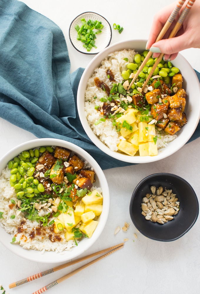 vegan teriyaki tofu bowls with pineapple, rice, and edamame. hand is using chopsticks.