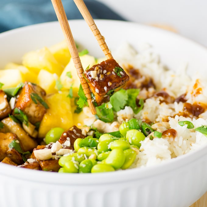 chopsticks holding teriyaki tofu over bowl with rice, pineapple, and edamame