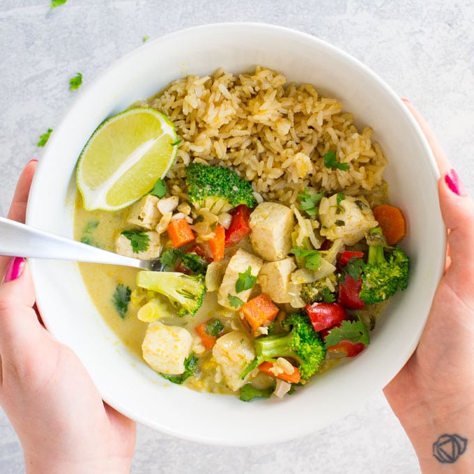 hands holding white bowl of vegan thai green curry with veggies, tofu, and brown rice