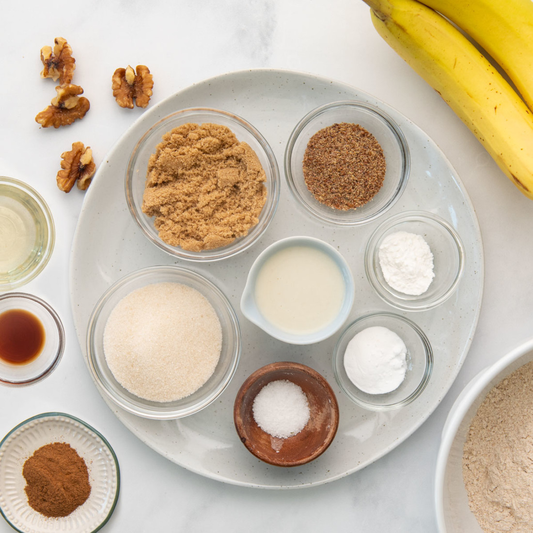 overhead view of sugar, brown sugar, almond milk, baking ingredients, bananas in measurement bowls on a large plate