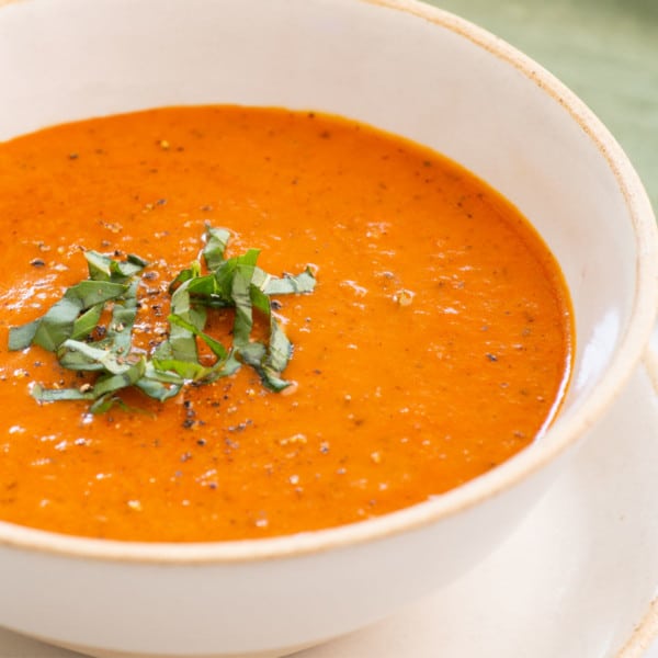 close up side view of a bowl of soup with tomato, carrots, aromatics and topped with fresh basil shreds