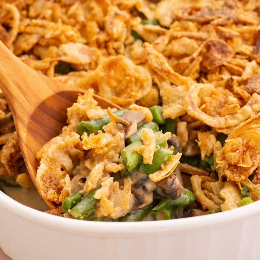 side view of wooden spoon scoop vegan green bean casserole with crispy fried onions in a white baking dish