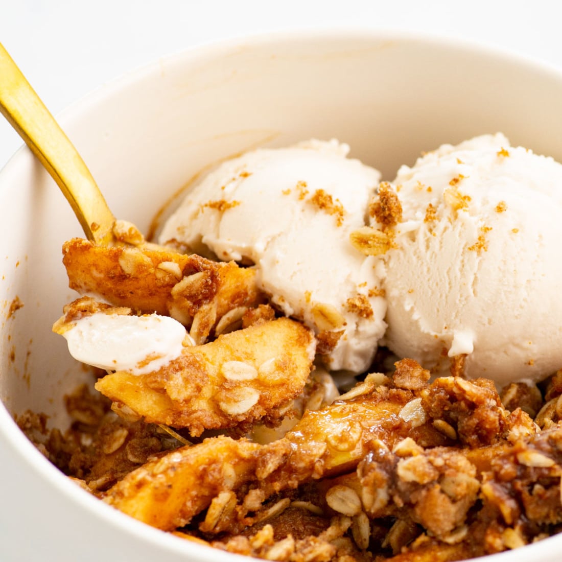 close view vegan apple crisp in a white bowl with two scoops of nondairy ice cream and gold spoon in the bowl