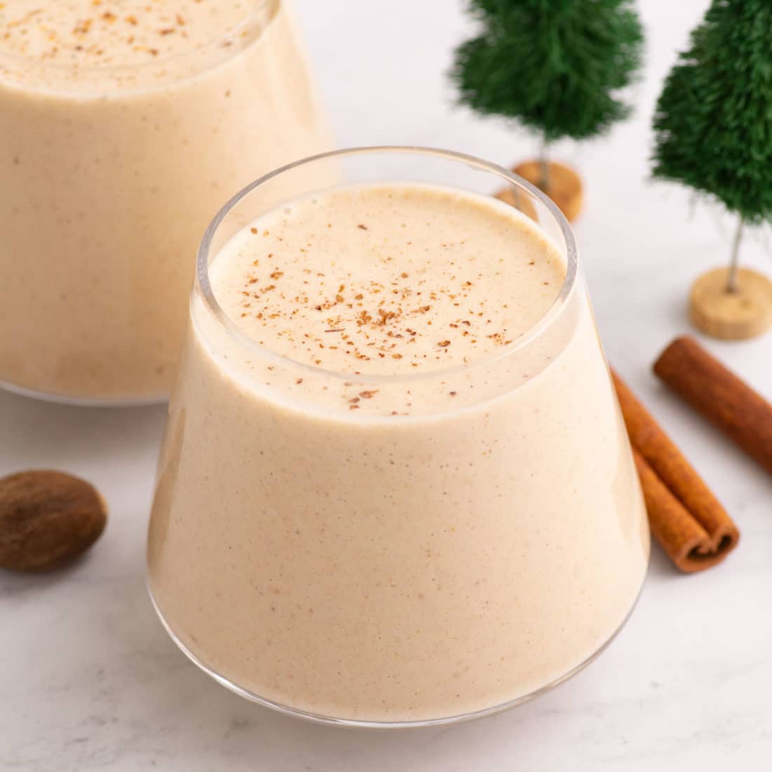eggnog in a glass with stick of cinnamon, nutmeg, and mini fake evergreen trees in background